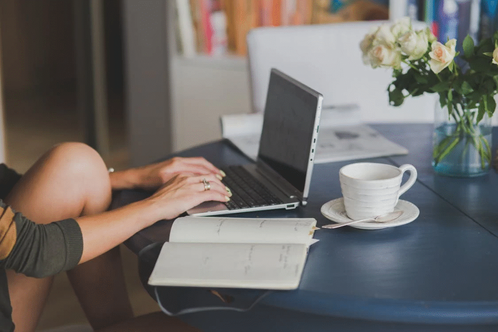 A person using a laptop on a table with a cup and a notebook
